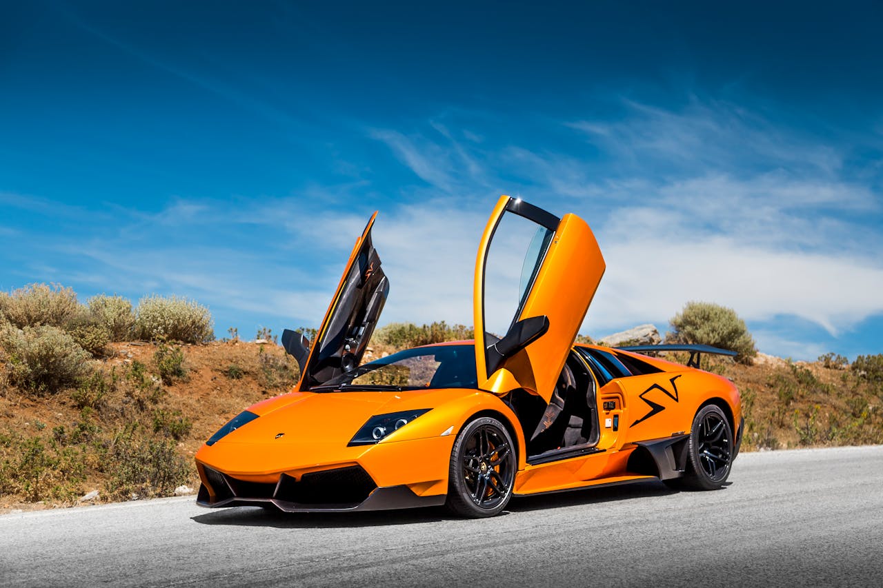 Orange sports car with doors open under bright blue sky on a scenic road.